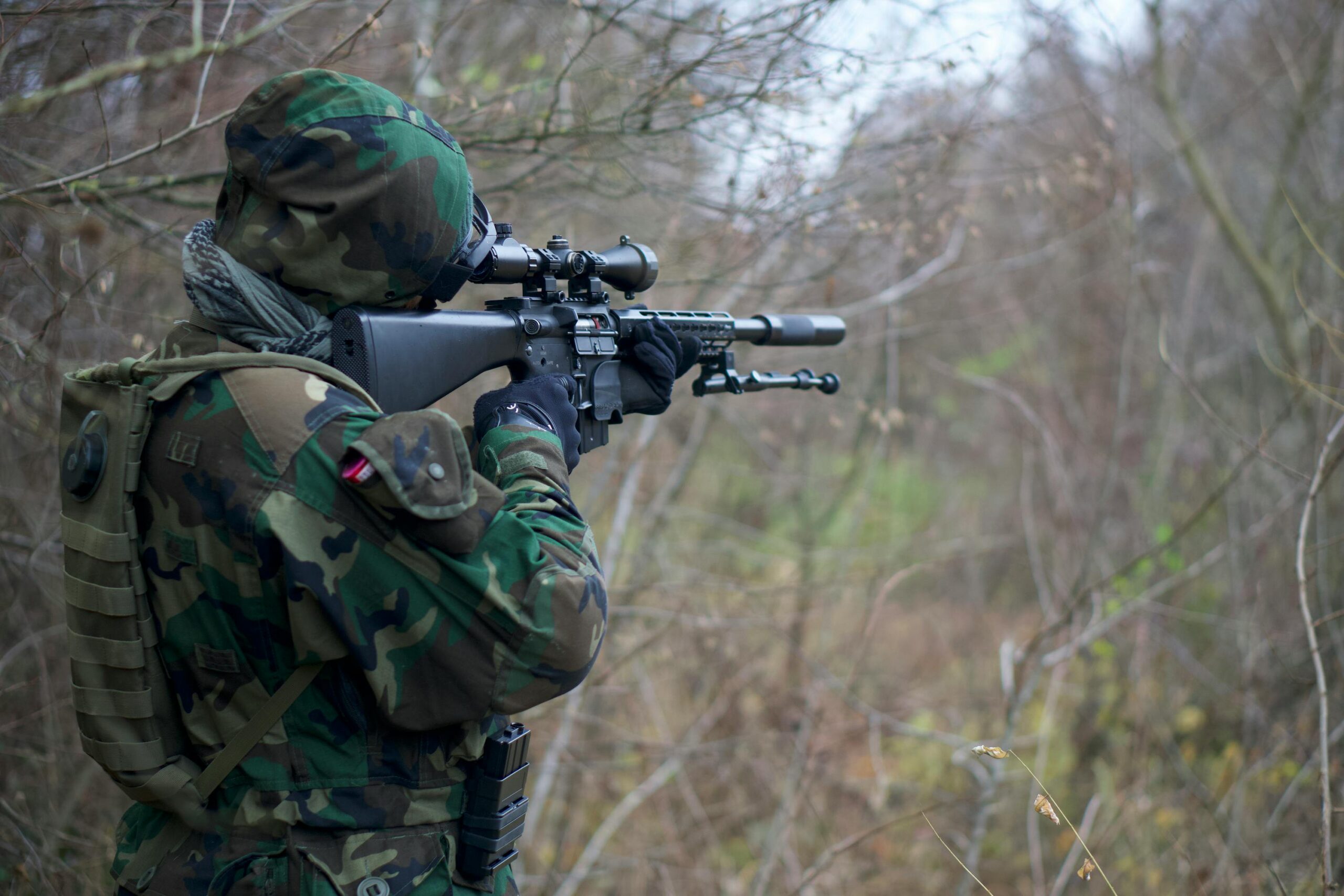Soldier in camouflage aiming a rifle in a dense forest setting during daylight.